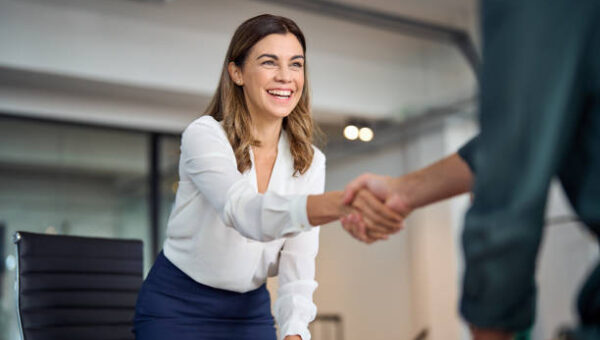 Happy mid aged business woman manager handshaking greeting client in office. Smiling female executive making successful deal with partner shaking hand at work standing at meeting table.